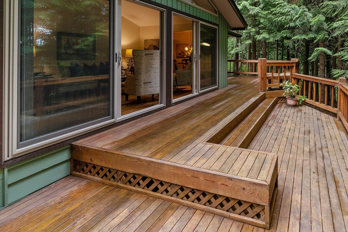 Cedar deck with a built-in bench and a wood railing behind a green house among tall evergreens