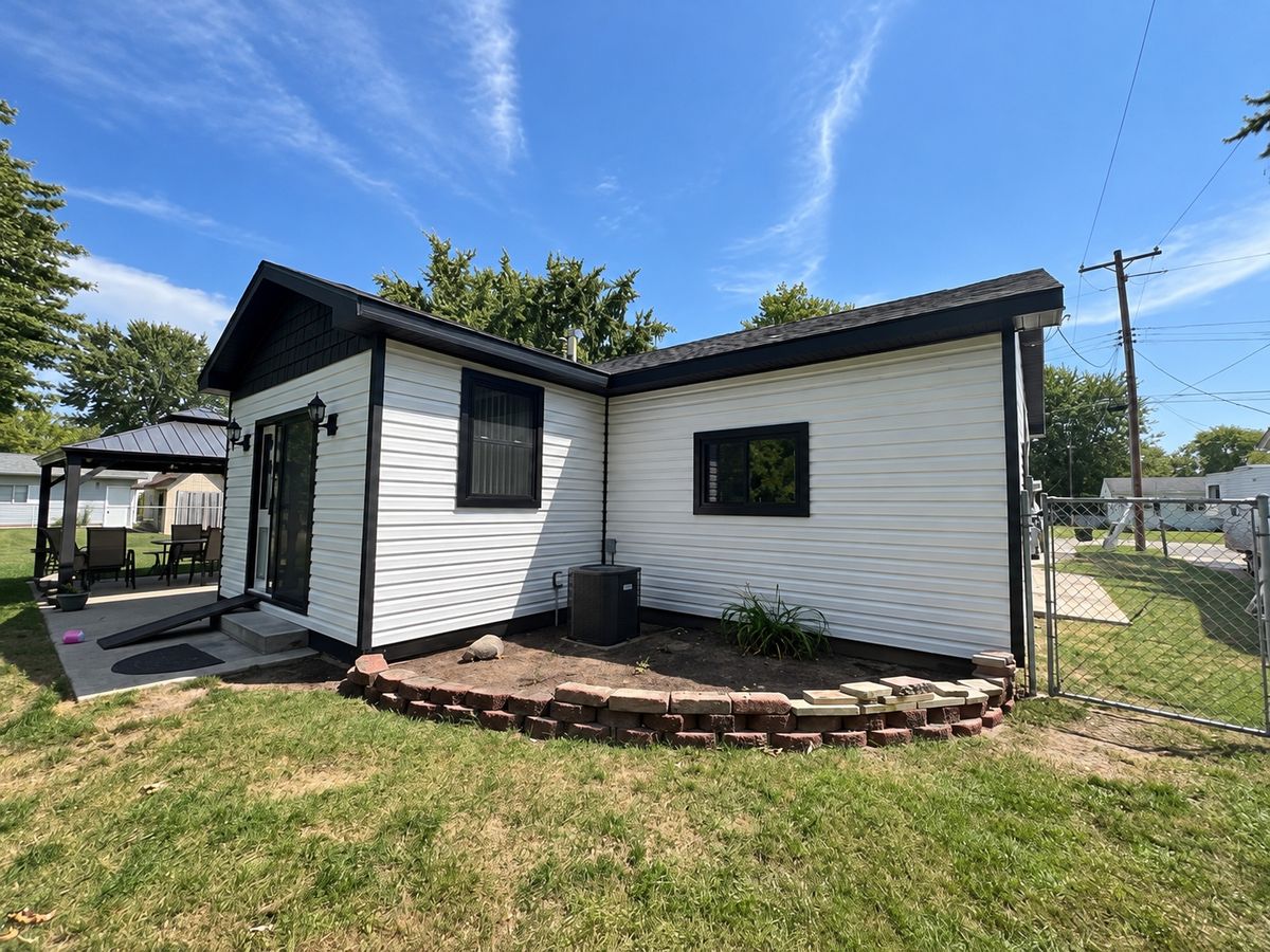 White horizontal vinyl siding on a small house with black trim, a glass door and a gazebo at the left