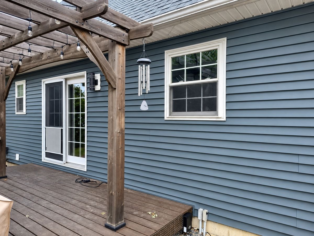 Rear wall of a house in slate blue-gray vinyl siding with a wood pergola over a brown deck and a double-hung window