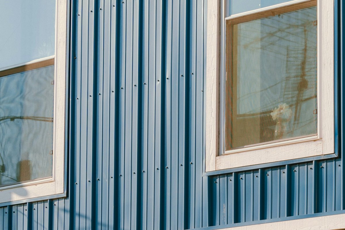 Blue vertical metal siding with exposed screws between two cream-trimmed windows
