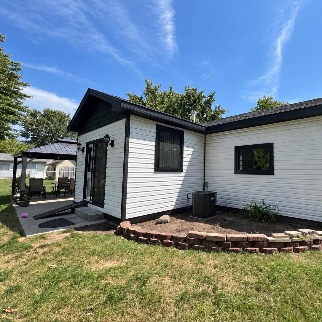 White horizontal vinyl siding on a small house with black trim, a glass door and a gazebo at the left