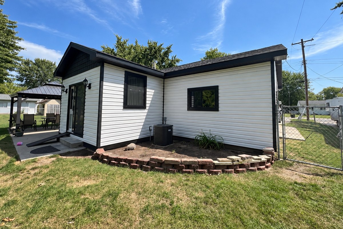White horizontal vinyl siding on a small house with black trim, a glass door and a gazebo at the left